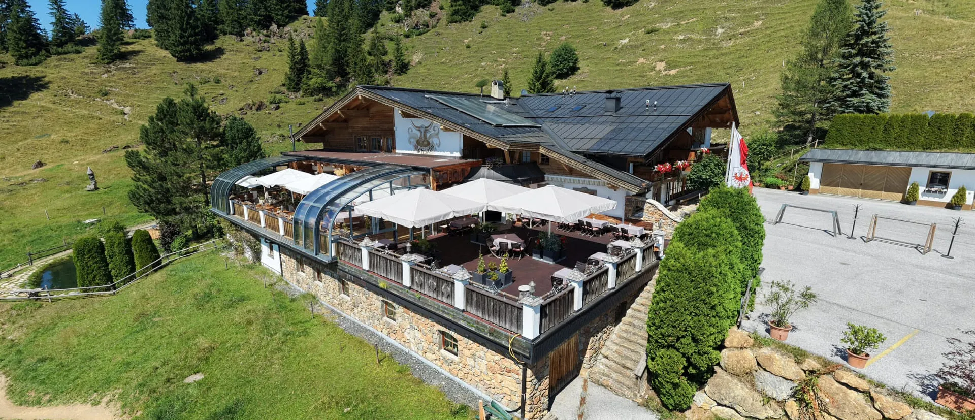 Drohnenaufnahme der OchsAlm: Sonnenterrasse mit weißen Sonnenschirmen, umgeben von Almwiesen und Bergwald